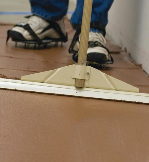 a man with spiked shoes in a bathroom using a squeegee to smooth a layer of brown Planicrete